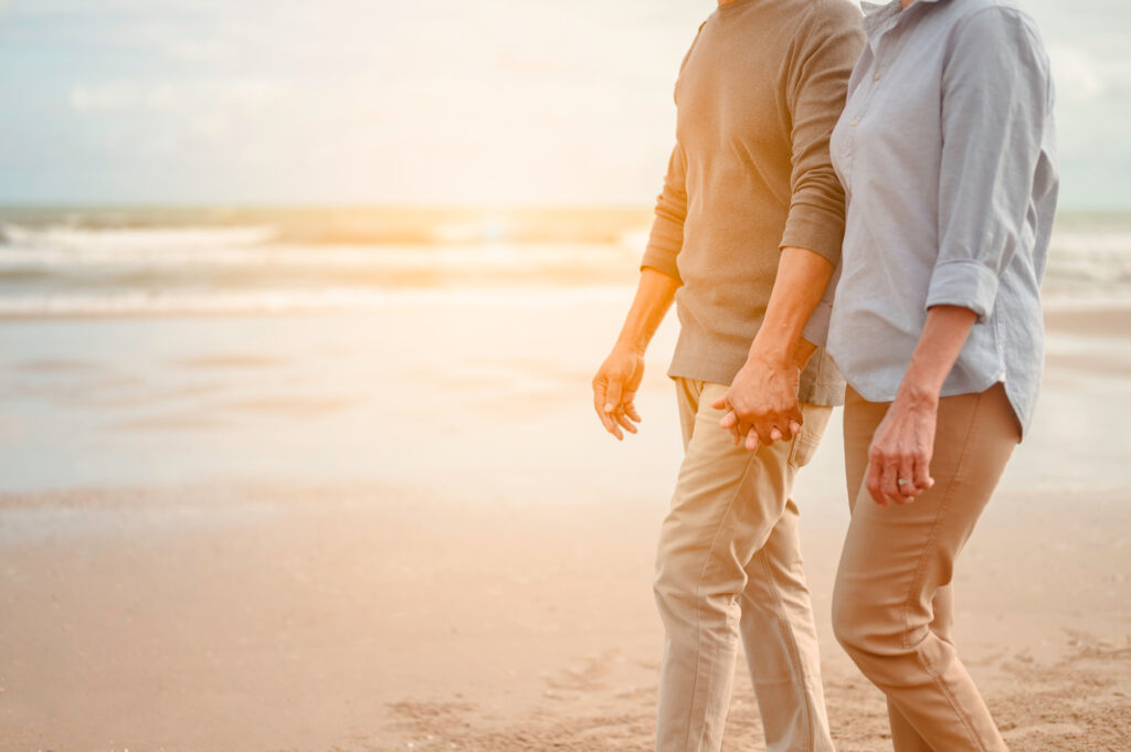 Senior lovers walk hand in hand at the beach at sunset.